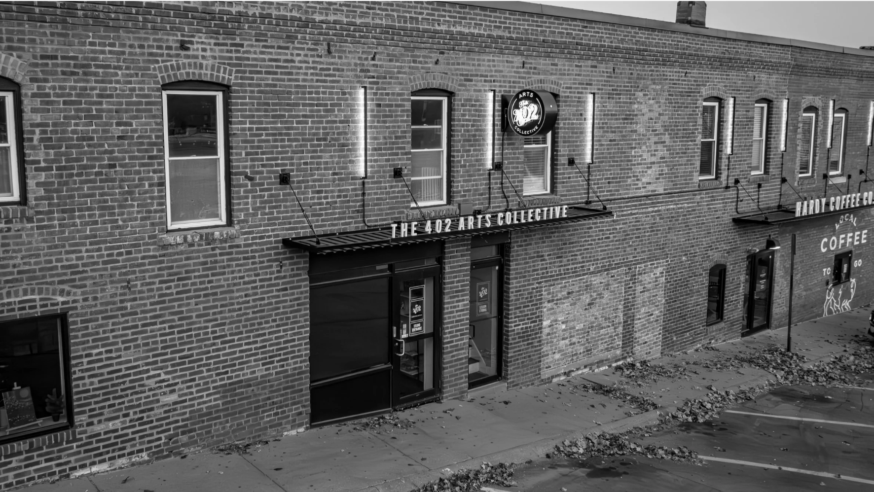 Black and white photo of a brick building with multiple illuminated windows and signs, including 'The 402 Arts Collective' and 'Hardy Coffee Co.' on the exterior.