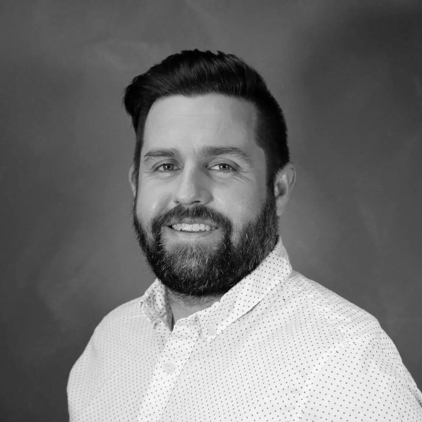 Black and white portrait of a man with dark hair, beard, and mustache, smiling, wearing a collared shirt with small polka dots, against a plain background.