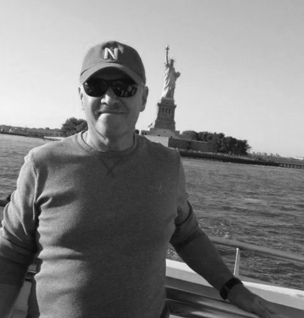 A man wearing sunglasses and a cap stands on a boat with the Statue of Liberty in the background.