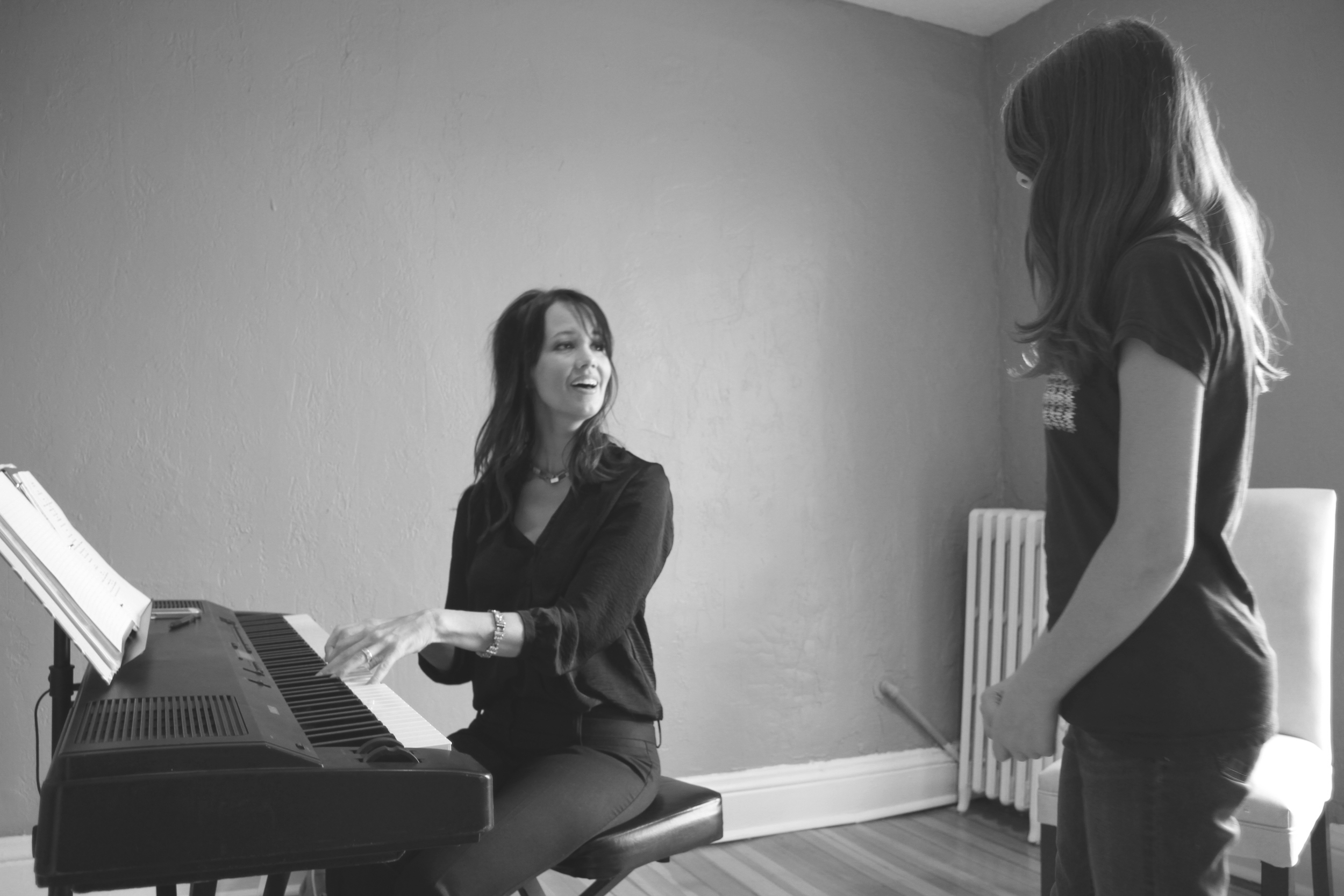 A woman playing a piano and smiling at a young girl standing in front of her in a room with plain walls and a radiator.