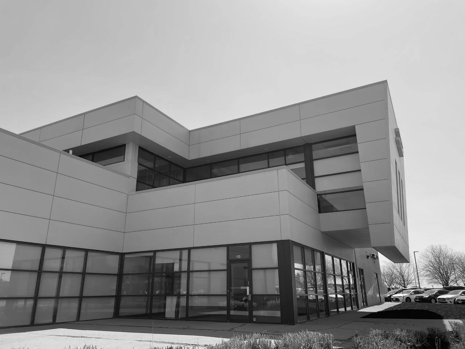 Modern commercial building with large glass windows and multiple stories, set against a clear sky with parked cars in the background.