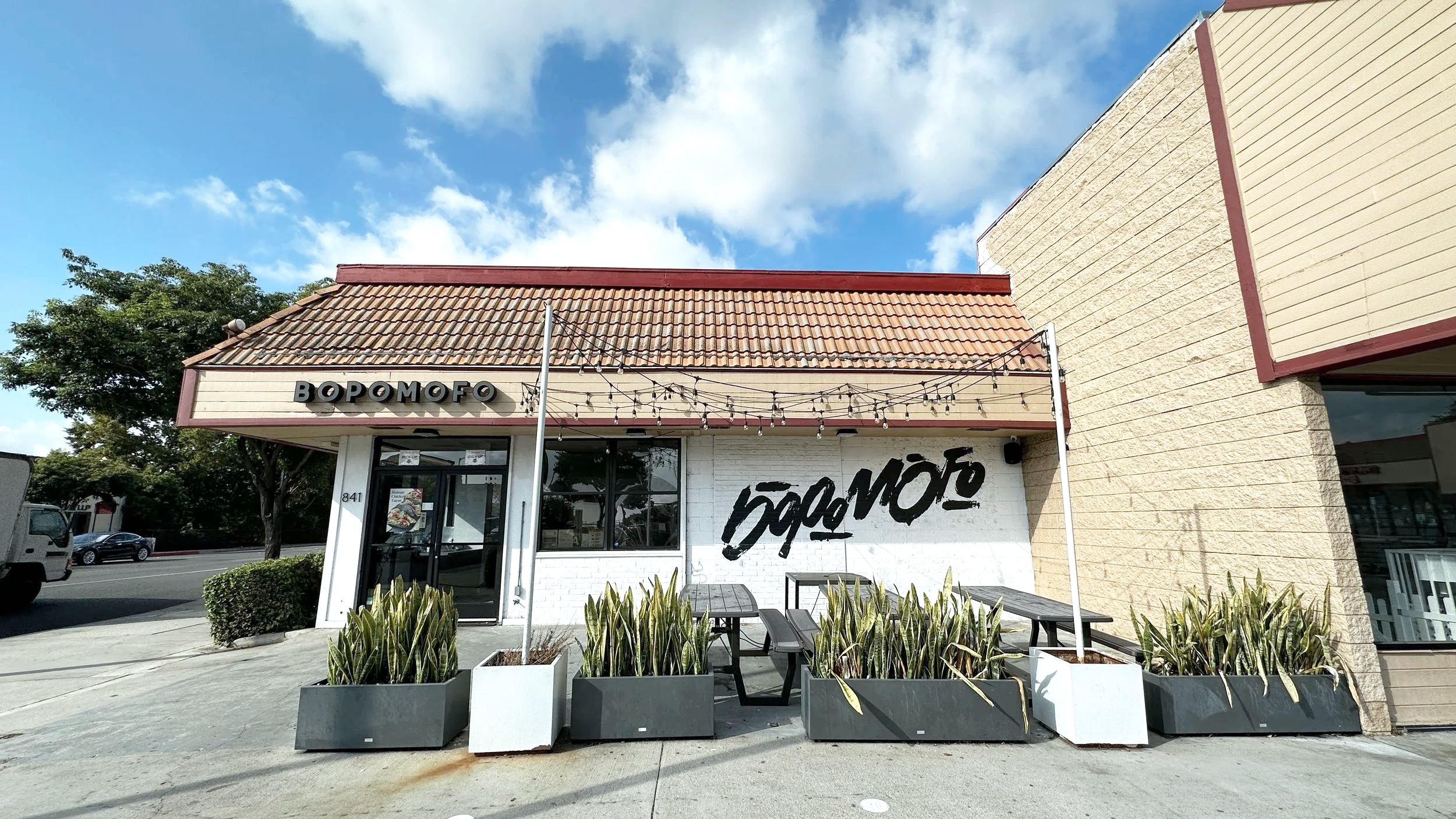 Exterior of a restaurant called BOPOMOFO with outdoor tables and planters, under a partly cloudy sky.