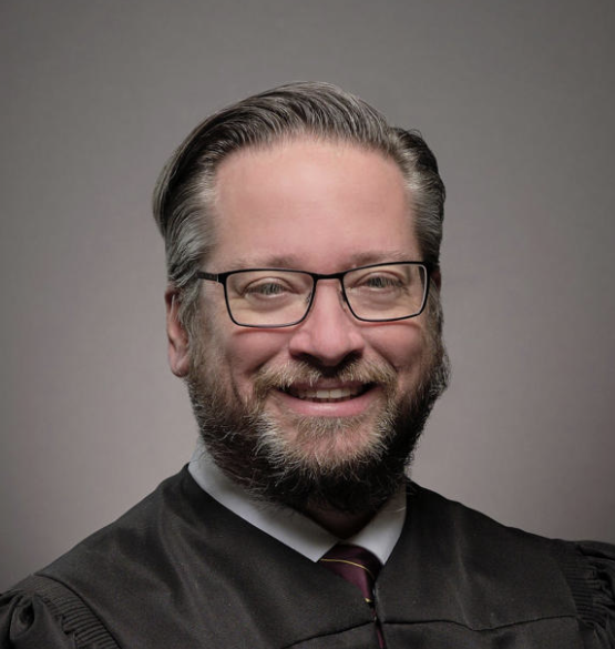 Portrait of a man with glasses, gray hair, and a beard, smiling in a black academic gown and tie against a gray background.