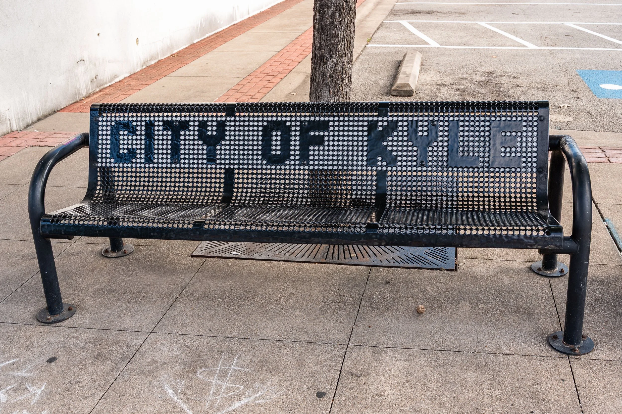 A metal bench with an opening in the backrest displaying the words 'CITY OF' and other letters indistinct, situated on a sidewalk near a parking lot with marked parking spaces and a tree.