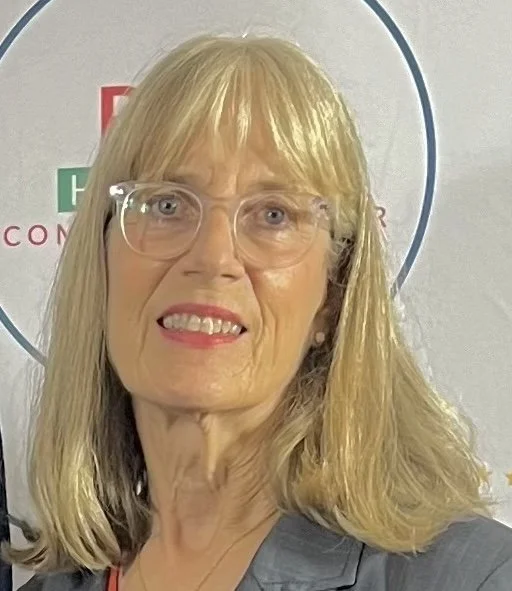 A woman with blonde hair, glasses, and a gray blazer standing in front of a gray wall with a partial logo visible.