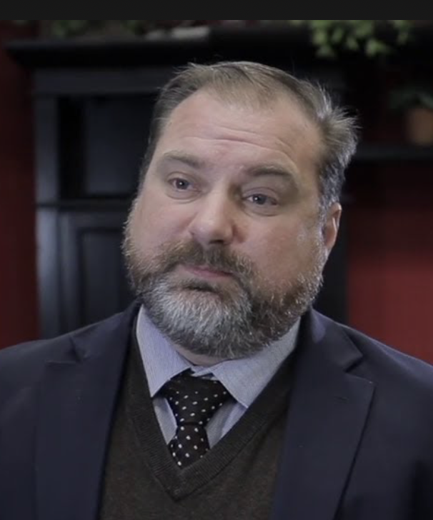 A middle-aged man with a beard and mustache, wearing a suit and tie, sitting indoors with a serious expression.