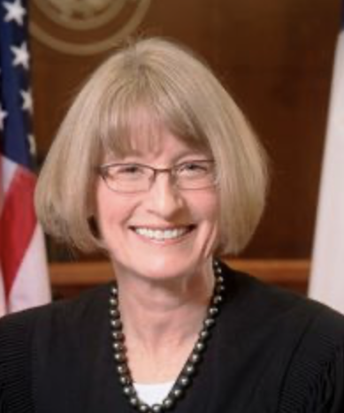 A woman with shoulder-length blond hair wearing glasses, a black outfit, and a pearl necklace, smiling in front of American flags.