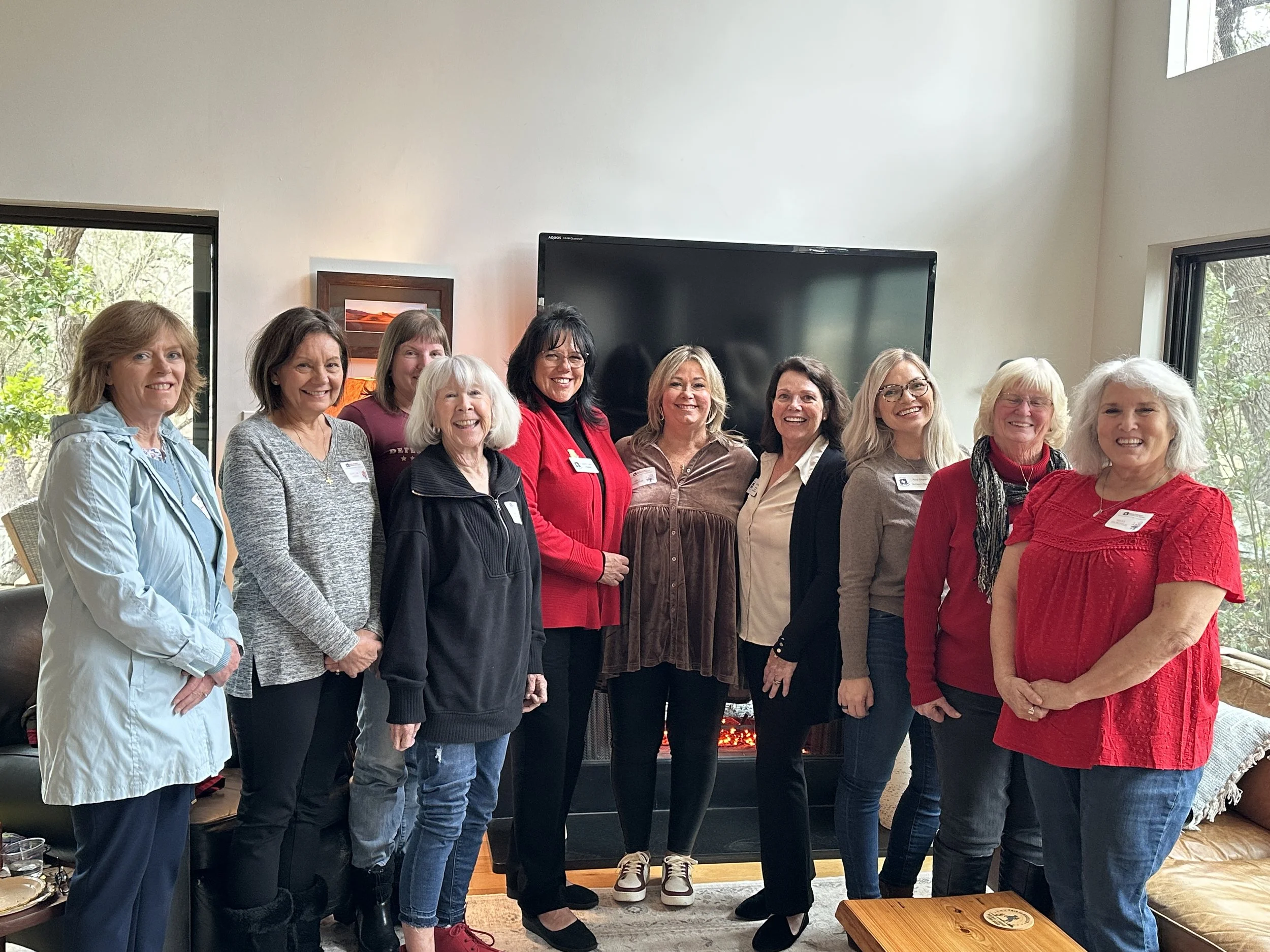Group of ten women standing indoors in front of a fireplace and a large TV, smiling at the camera.