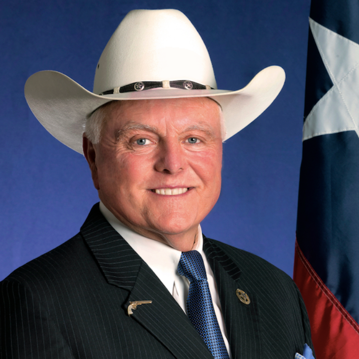 A man in a dark pinstripe suit, white shirt, and blue tie, wearing a white cowboy hat, standing in front of a Texas flag with a blue background.