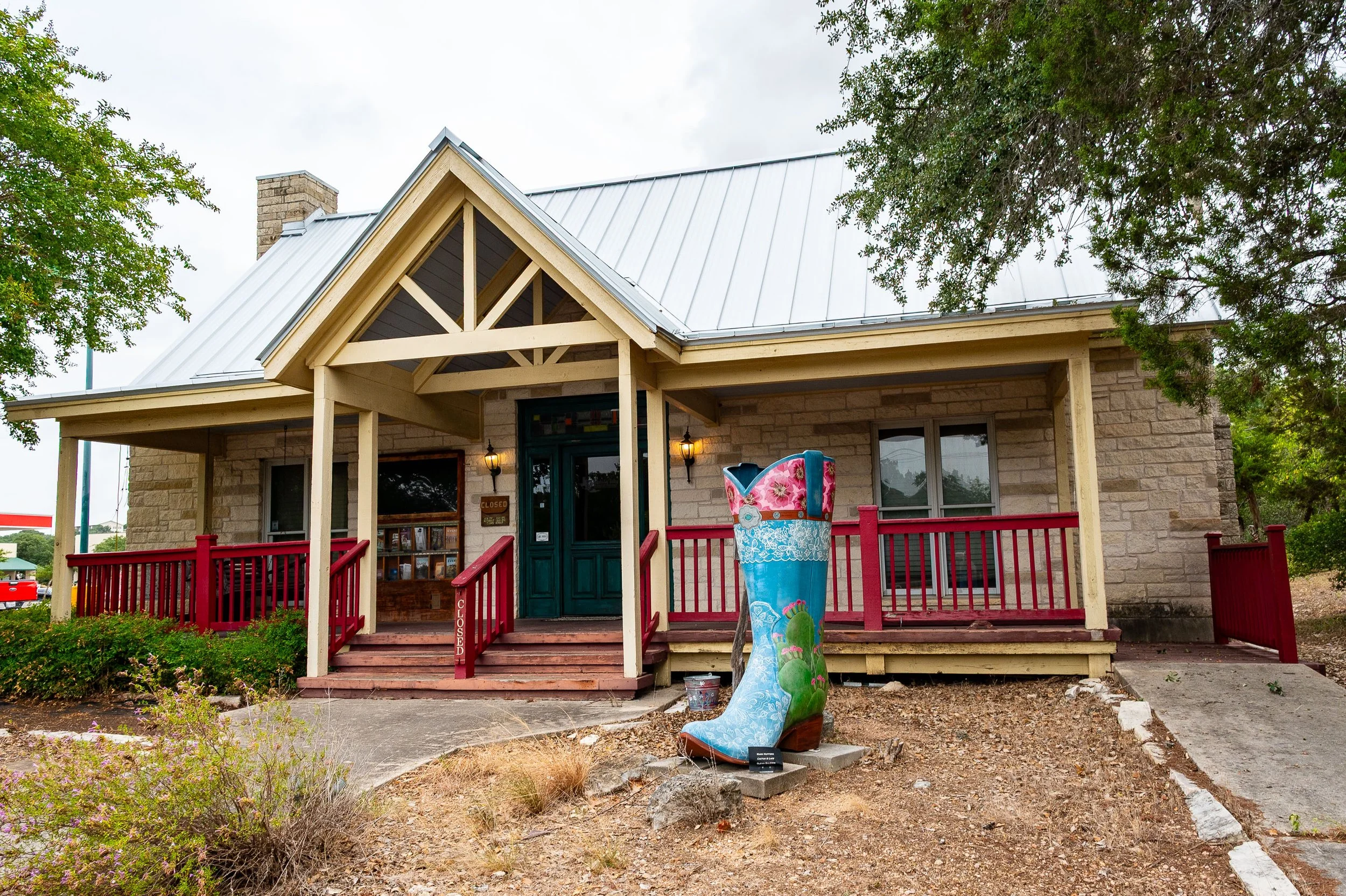 A house with a porch and red railing, with a large painted cowboy boot sculpture in front, surrounded by natural landscaping.