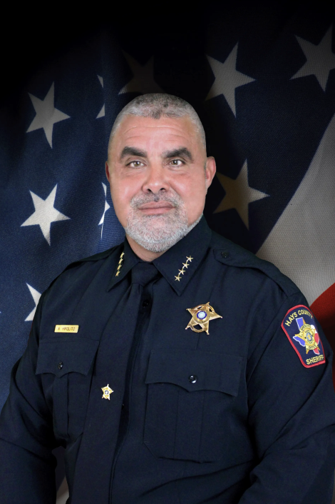 Portrait of a male police officer in uniform in front of an American flag backdrop.