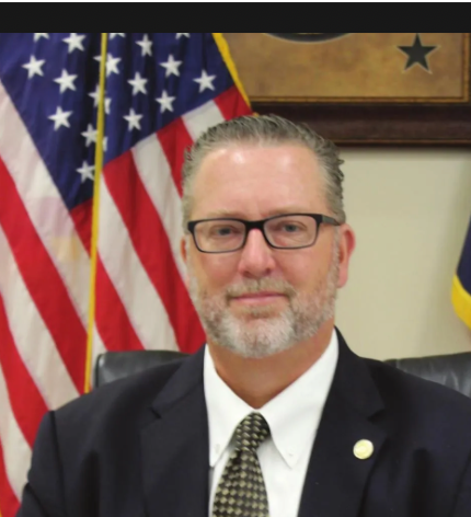 A man with glasses and a beard, wearing a suit and tie, sitting in front of an American flag and a Texas flag in an official setting.