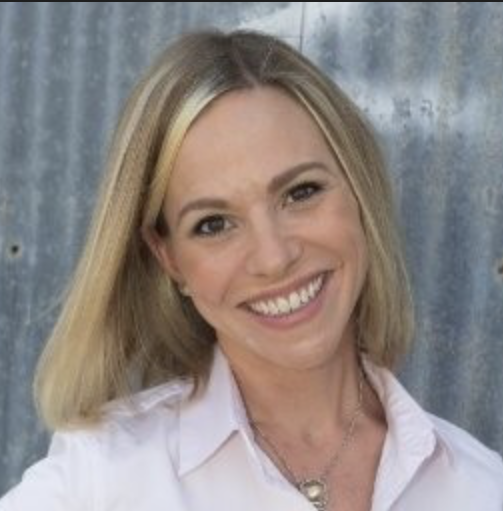 A smiling blonde woman wearing a white shirt in front of a metallic backdrop.