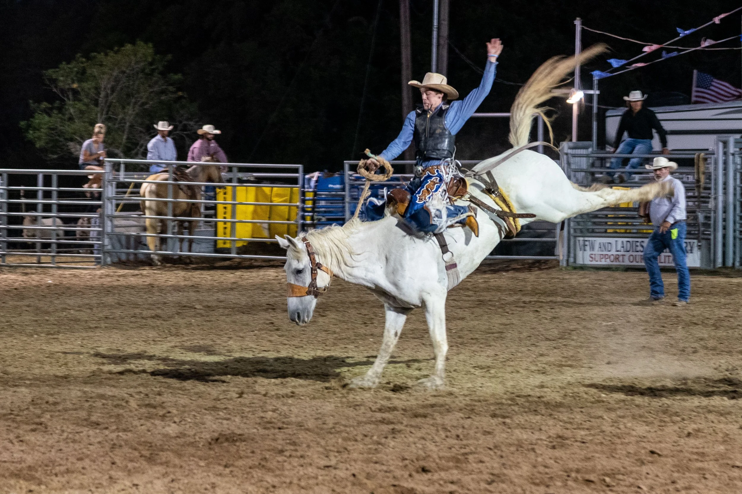 A cowboy riding a bucking white horse at a rodeo, with spectators and other cowboys on horseback in the background.