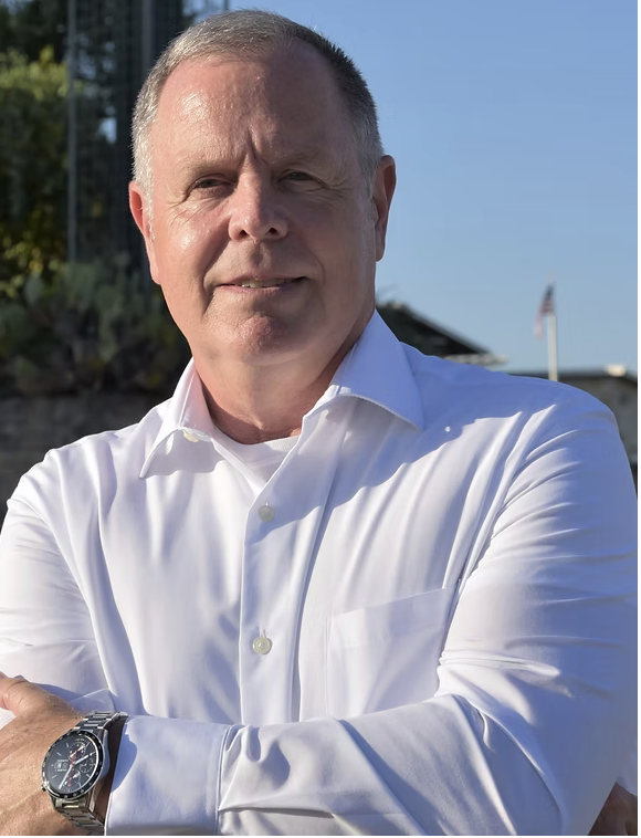 A middle-aged man with short gray hair wearing a white dress shirt, standing outdoors with a blue sky, some greenery, and a building with an American flag in the background.
