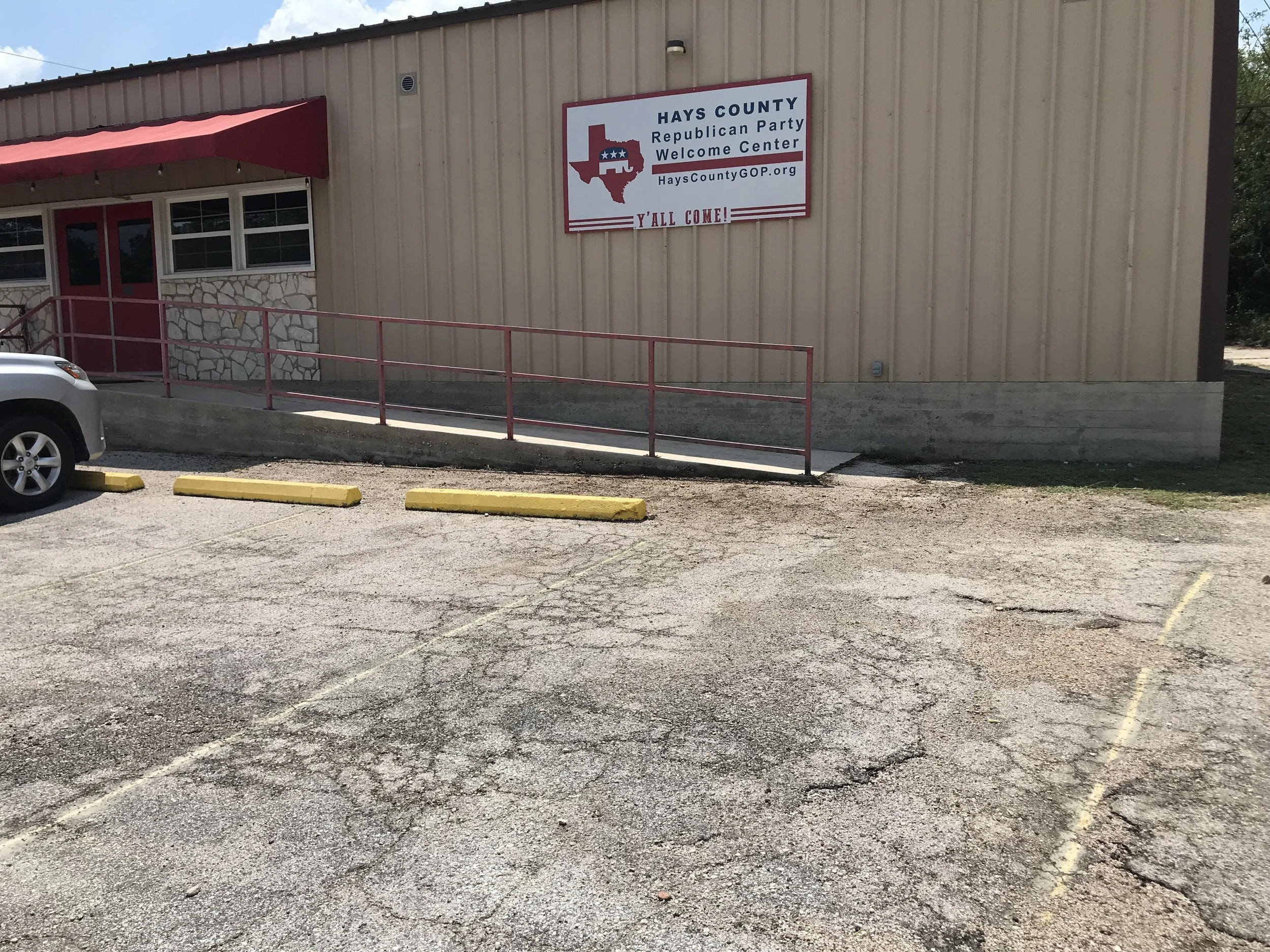 Building with a sign that reads, 'Hays County Republican Party Welcome Center,' with a Texas map outline and below it, 'Y'all Come!' There are yellow parking blocks and a ramp with red railings in the foreground.