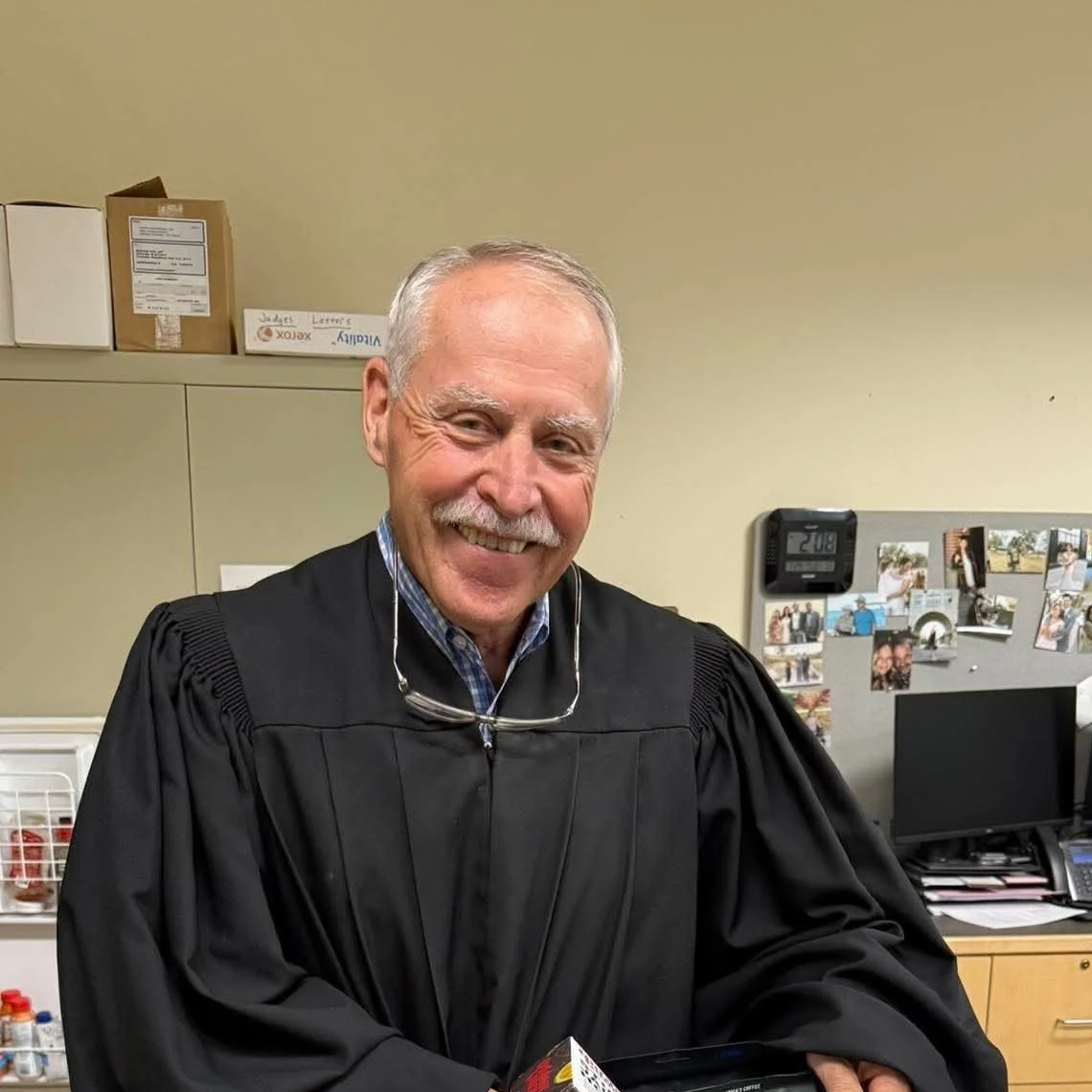 Older man in a black judicial robe smiling in an office setting.