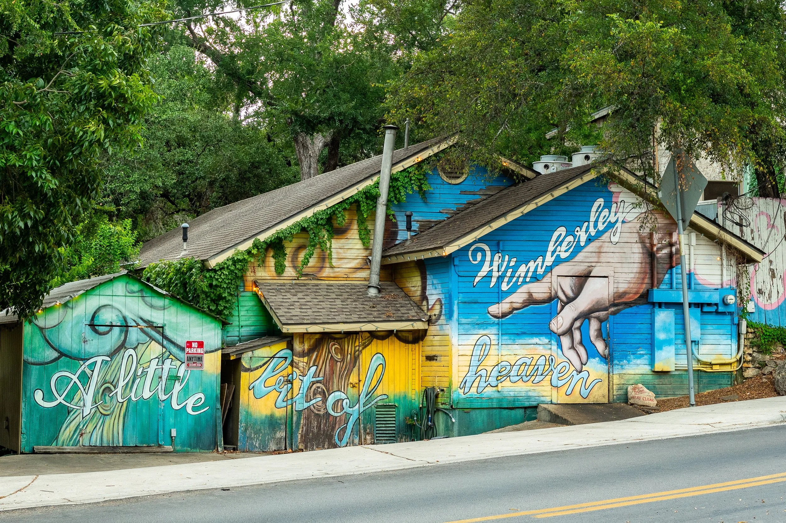 Colorful mural on the side of a building with trees in the background. The mural features the words 'A little to get al our hearts' and a large hand pointing. The building has gray shingles and various pipes on the roof.