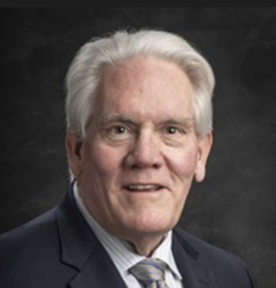Portrait of an older man with white hair, wearing a dark suit, striped shirt, and patterned tie, against a dark background.