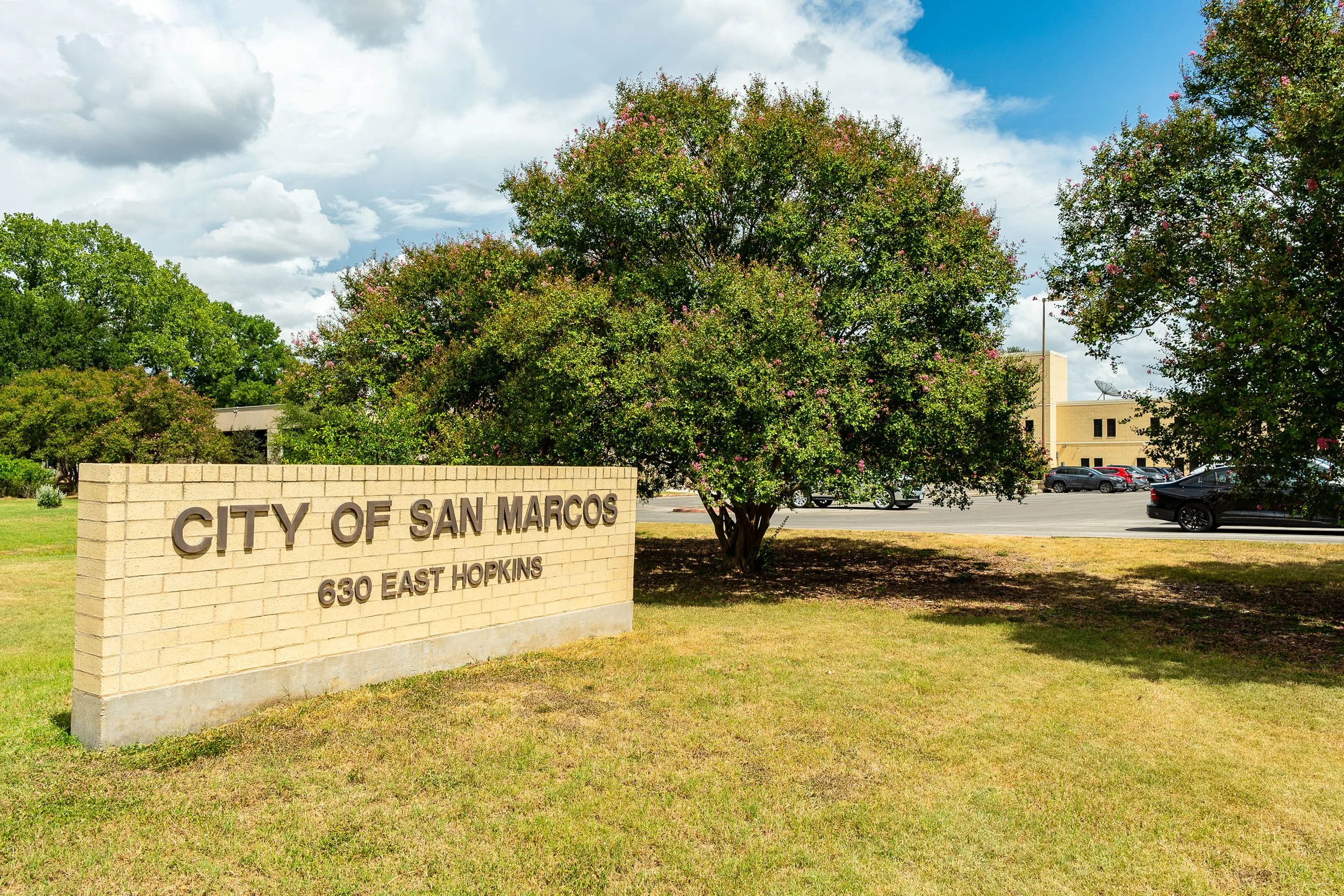 A brick sign for the City of San Marcos with the address 630 East Hopkins, situated in front of a large green tree on a grassy area. In the background, there is a parking lot and a beige building under a partly cloudy sky.