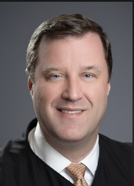 Professional headshot of a man in a suit and tie with a gray background.