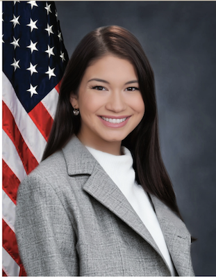 A young woman with long dark hair smiling, wearing a gray blazer and white shirt, standing in front of an American flag.
