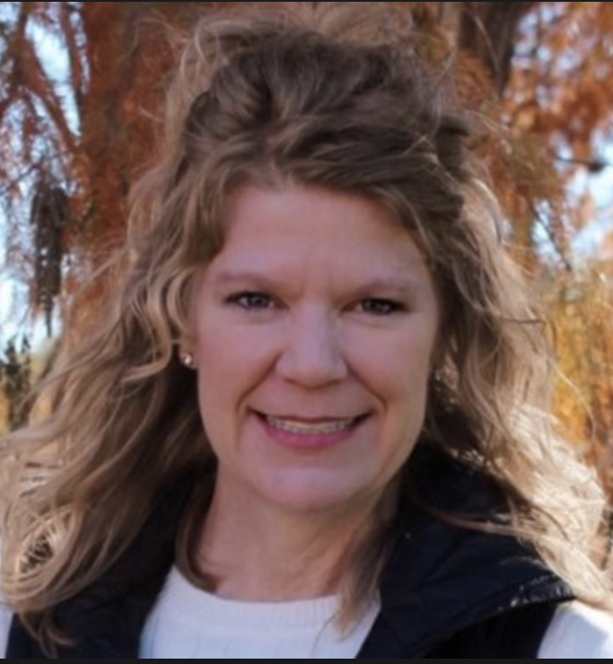 A woman with curly blonde hair smiling outdoors with autumn leaves in the background