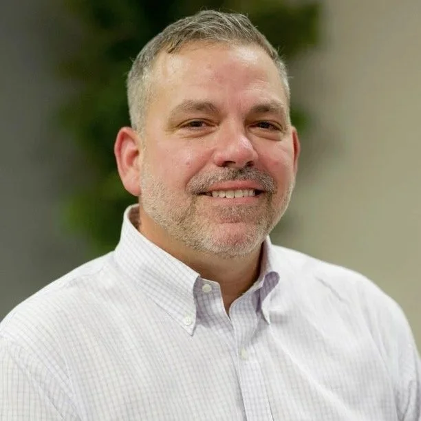 A middle-aged man with short gray hair and a beard, smiling, wearing a white button-up shirt with a checkered pattern, standing in front of a blurred background of greenery.
