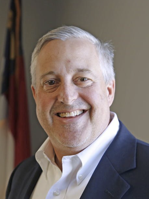 Portrait of a smiling middle-aged man with short gray hair, wearing a white shirt and dark blazer, standing indoors with curtains in the background.