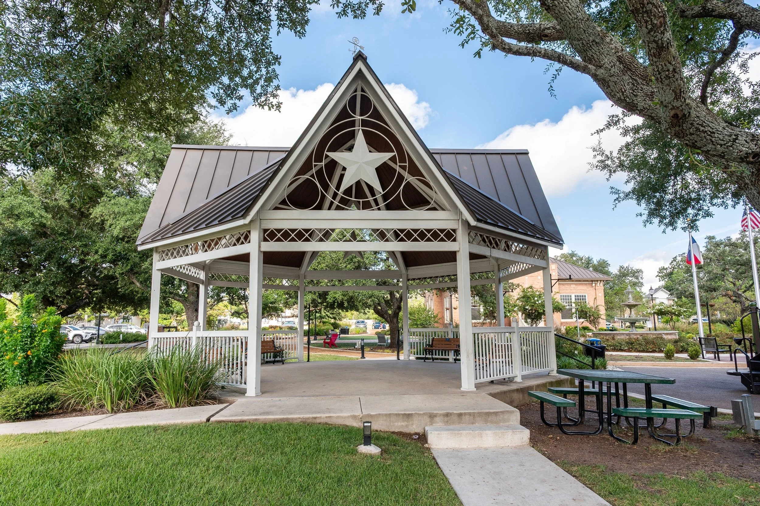 A park gazebo with a star and circle design on top, surrounded by trees, grass, chairs, and tables.