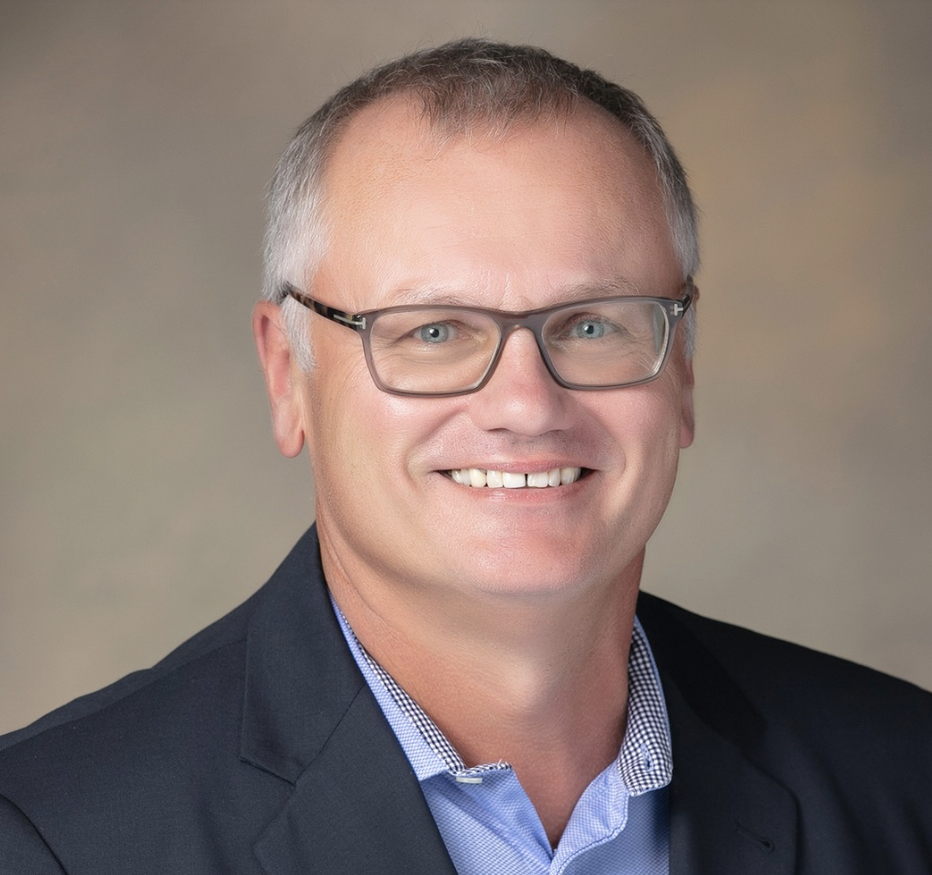 Headshot of a smiling middle-aged man with short gray hair, glasses, wearing a dark blazer and a collared shirt, against a neutral background.