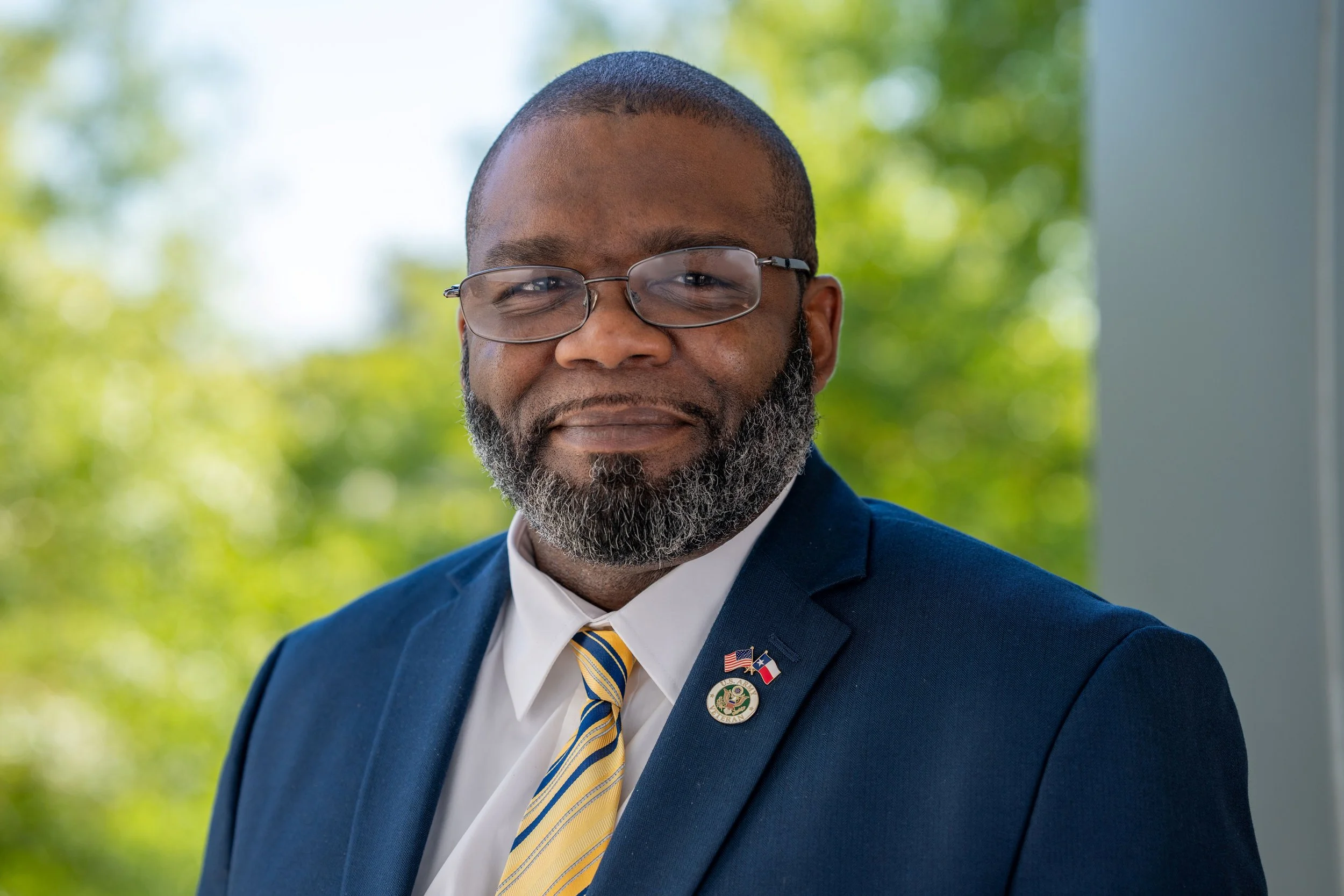 A man with glasses, a beard, and short hair, wearing a navy suit with American and Texas flag pins, a white shirt, and a yellow striped tie, standing outdoors with blurred green trees in the background.