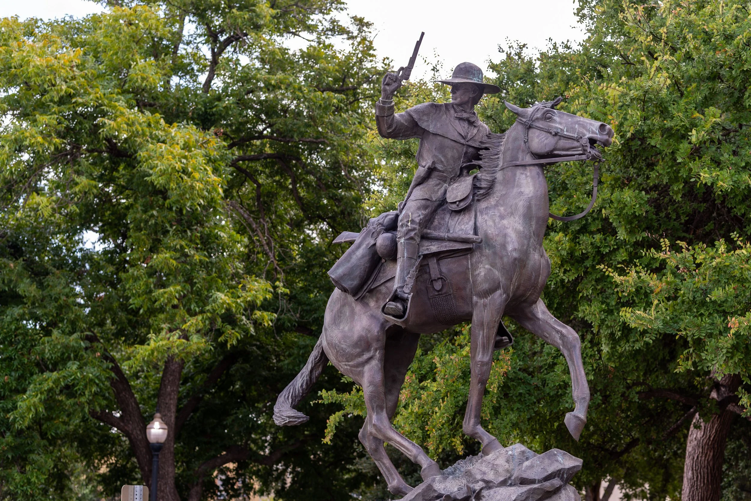 Bronze equestrian statue of a man on a horse, holding a rifle, with trees in the background.