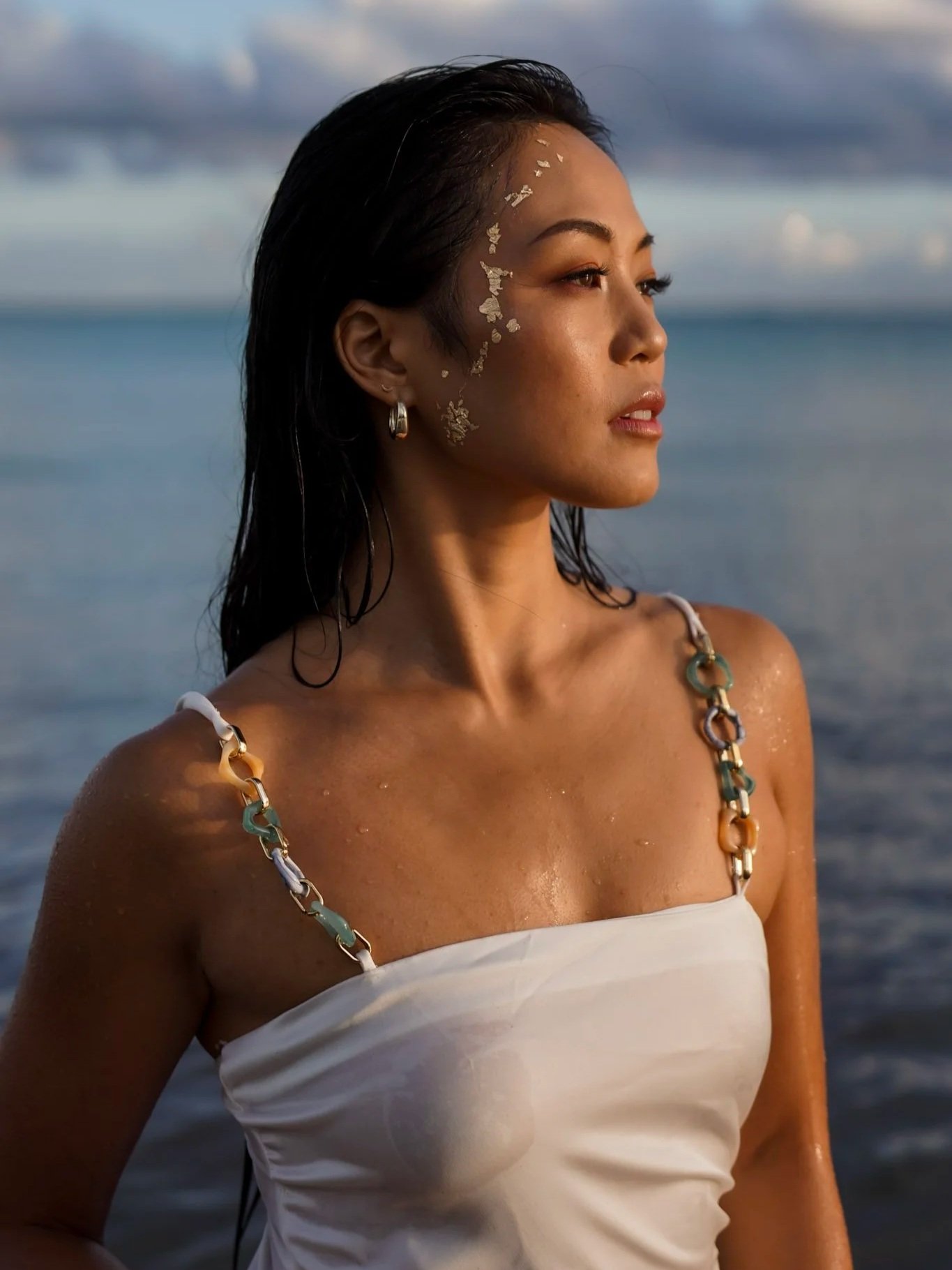 A woman with dark hair and gold face decoration standing by the ocean, wearing a white tube top with chain straps.