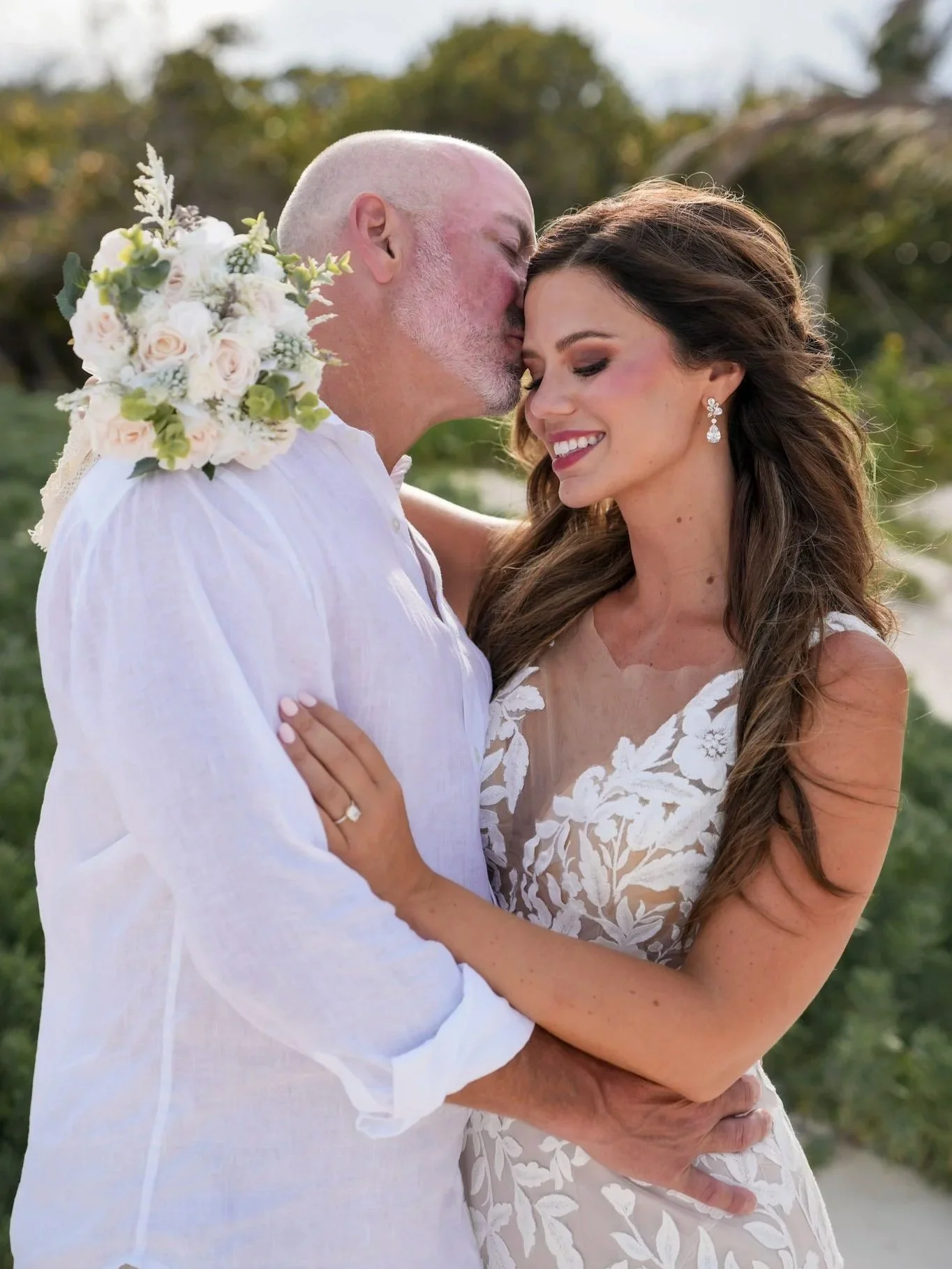 A couple, dressed in wedding attire, embracing outdoors on a sunny day, with the man kissing the woman on her forehead.