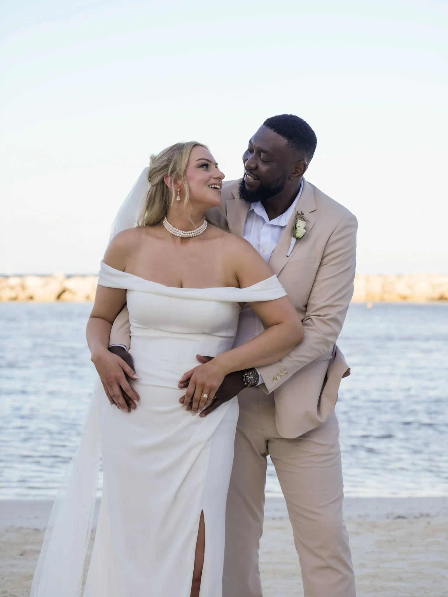 A couple dressed in wedding attire, standing on a beach with water and rocks in the background, smiling and looking at each other.