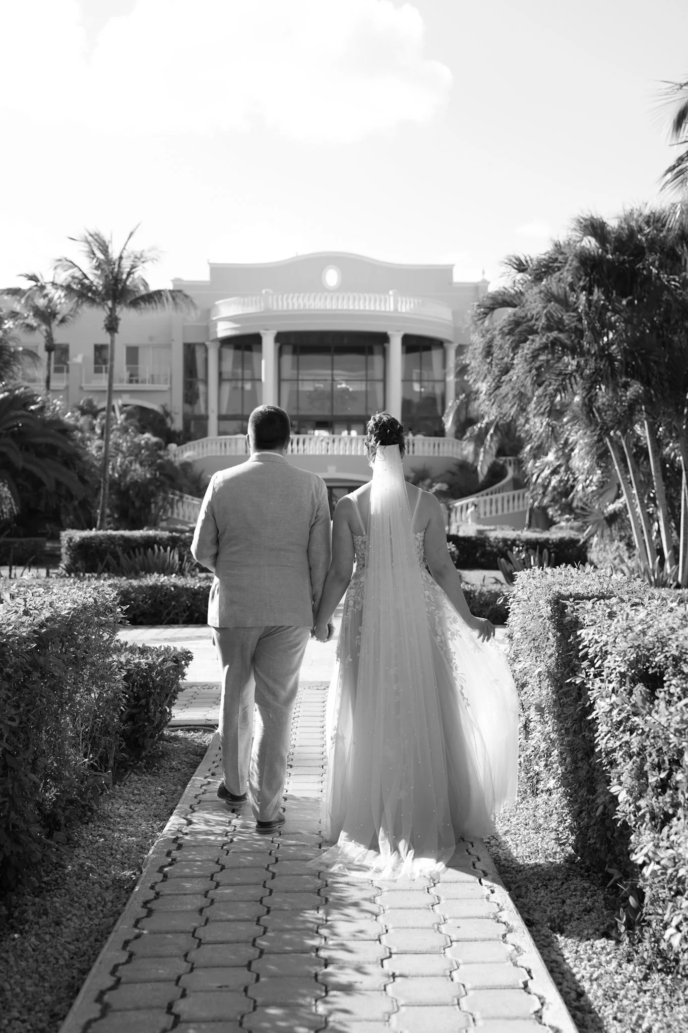 A couple, a man in a suit and a woman in a wedding gown, walk hand in hand down a paved garden path towards a large house, with palm trees and lush bushes on either side, under a clear sky.