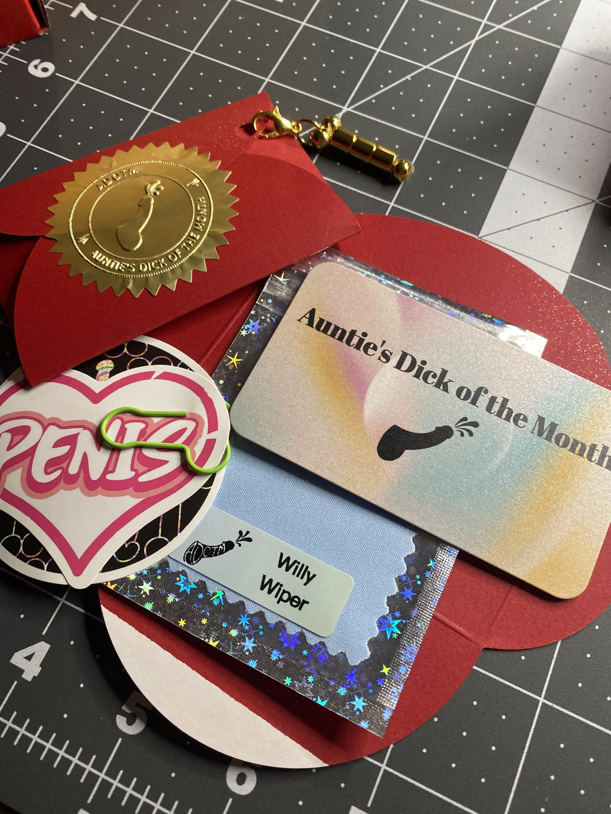 Red envelope with decorative stickers and cards, including one labeled "Auntie's Dick of the Month" with a cartoon of a penis, a "PEN15" sticker with a paperclip, and a gold tare weight with a clip, on a cutting mat.