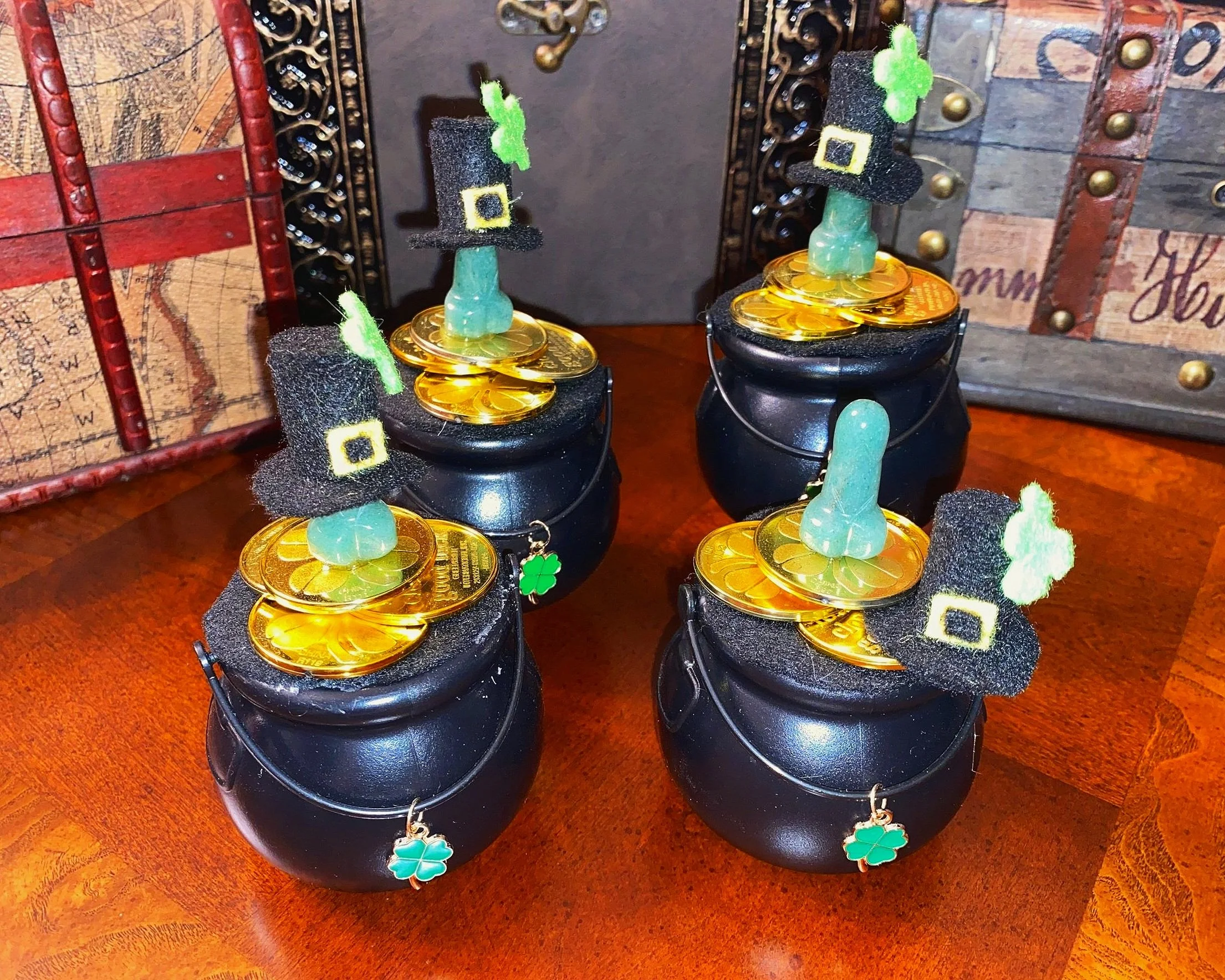 Four black pots filled with gold Dollar coins, decorated with small green shamrocks, miniature black top hats with green shamrocks, and small green clover charms, arranged on a wooden surface with a decorative wooden box and framed map in the background, indicating a St. Patrick's Day theme.