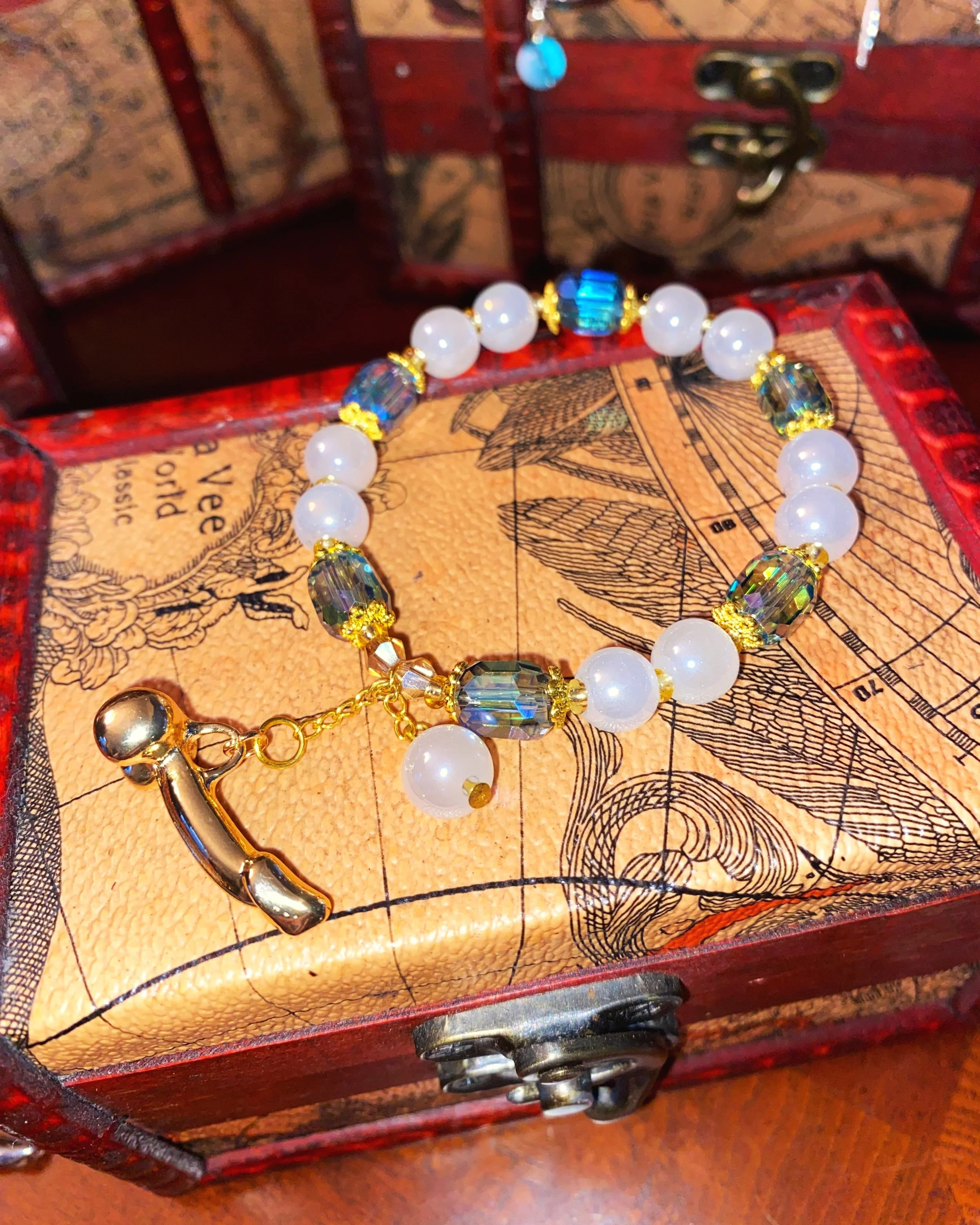 A vintage-style bracelet with white pearls, faceted crystal beads, and a small gold charm in the shape of a telephone, displayed in an antique jewelry box with a map design.