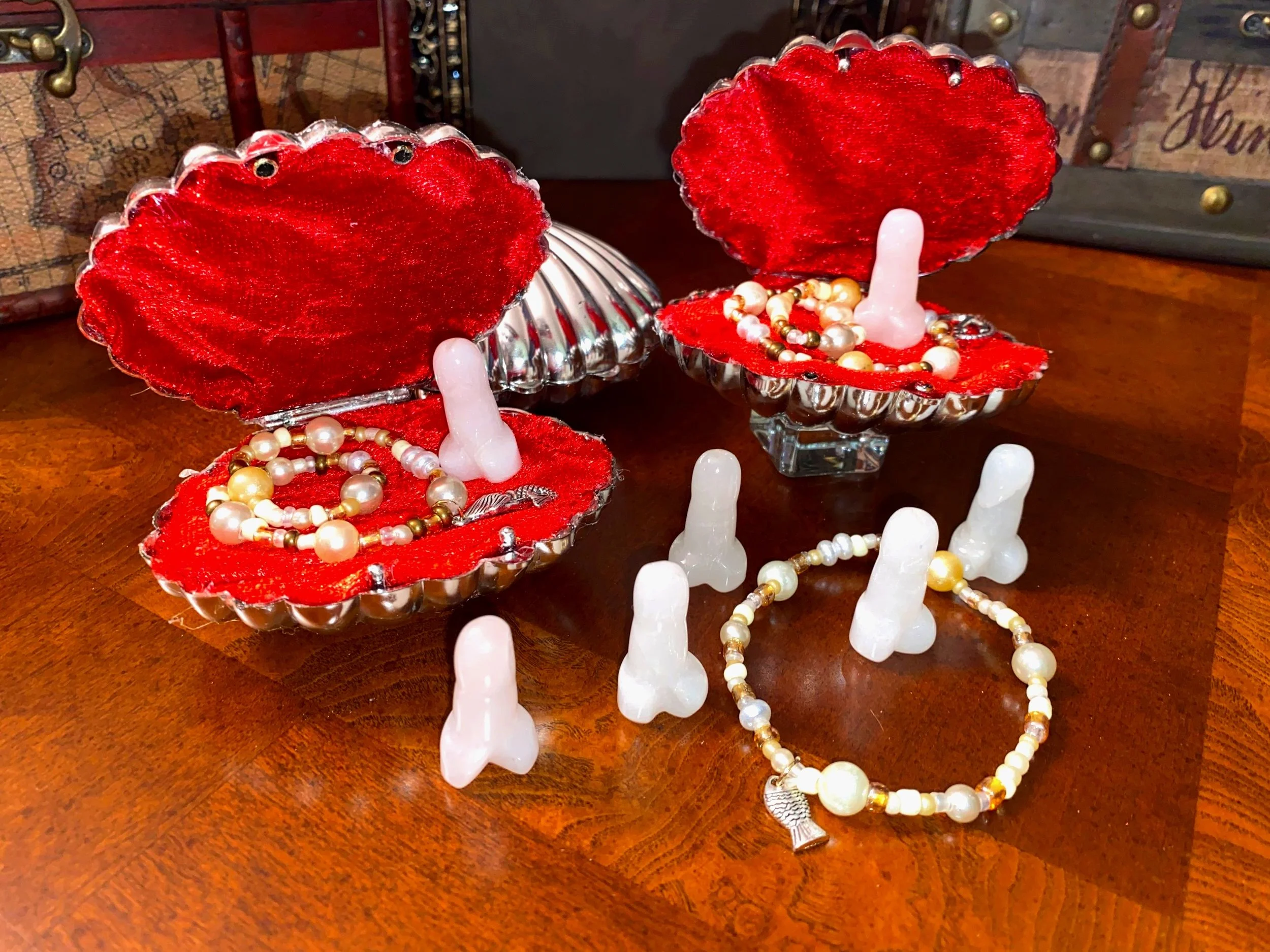 Open red velvet jewelry boxes with pearl necklaces and bracelets, surrounded by white ceramic penis-shaped objects, on a wooden table.