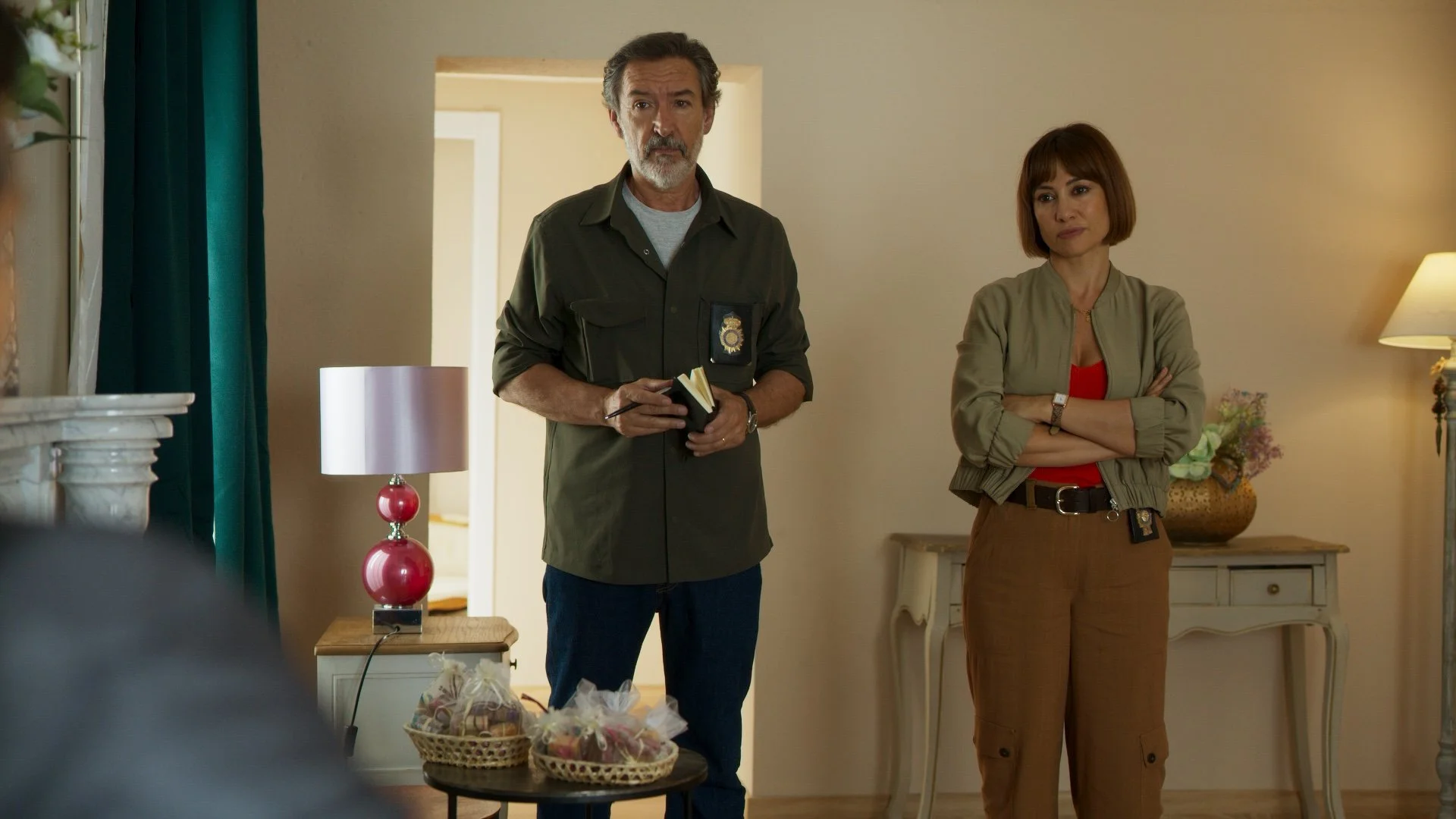 Una vida menos en Canarias Netflix - Two people standing in a living room with a notepad and a badge, wearing casual attire; a lamp and decorative items are in the background.