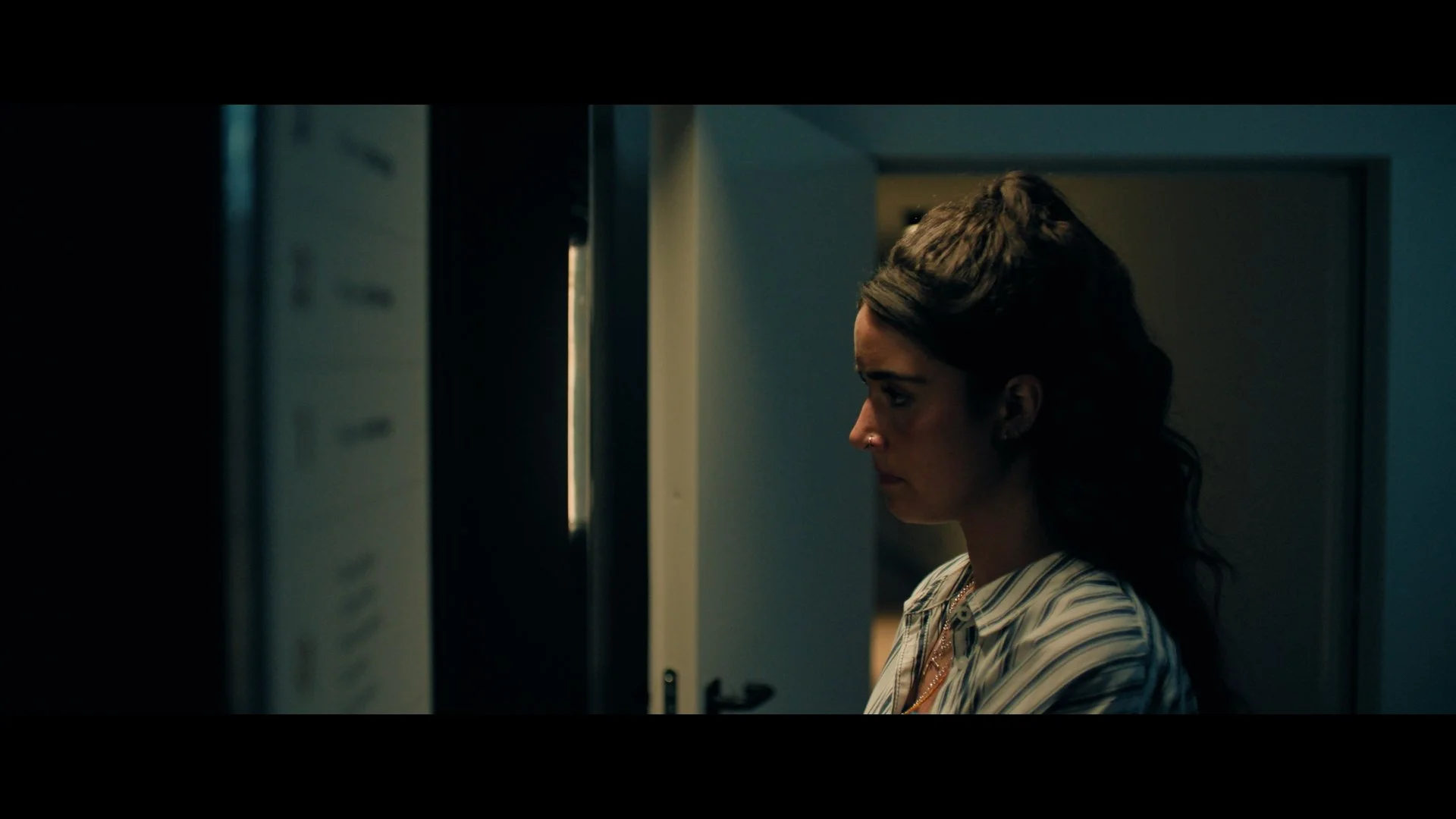 my fault London film A woman with dark hair in a striped shirt standing indoors, looking towards a partially open door.