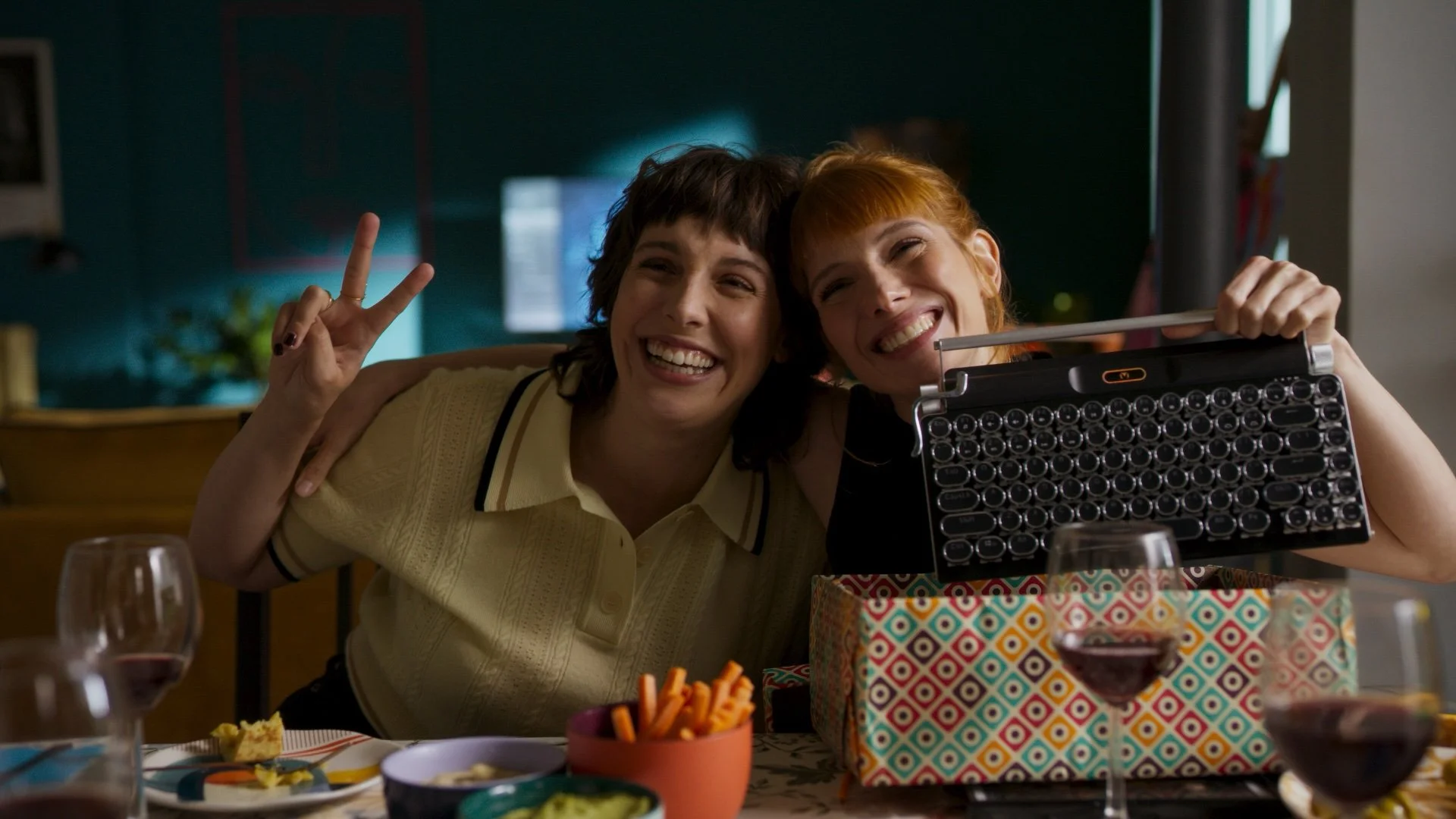Valeria Netflix temporada 4 Two women smiling and posing at a table with wine glasses, one making a peace sign, the other holding a typewriter-like device, with snacks on the table.