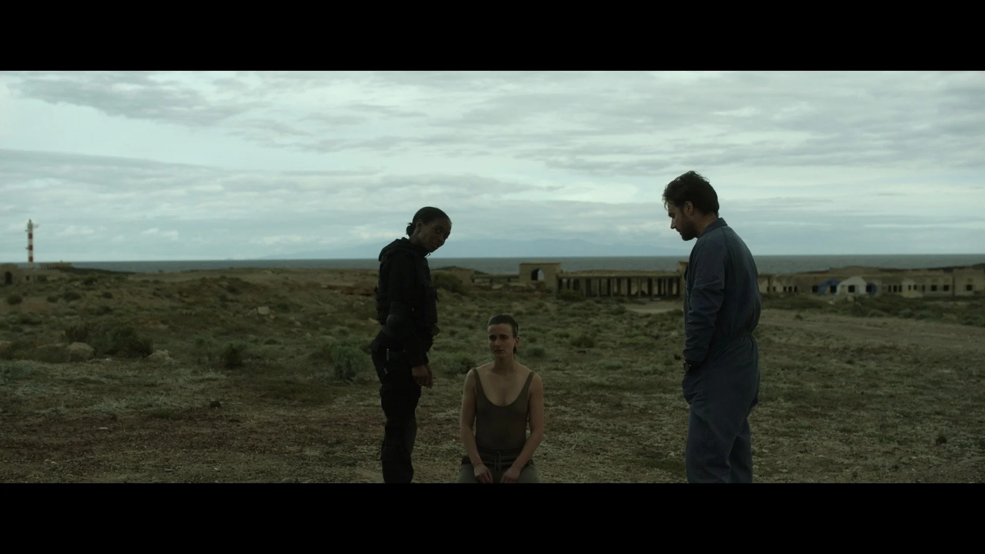 Three people in a desolate outdoor setting, with two standing and one kneeling. A barren landscape with sparse vegetation and old buildings in the background, under a cloudy sky.