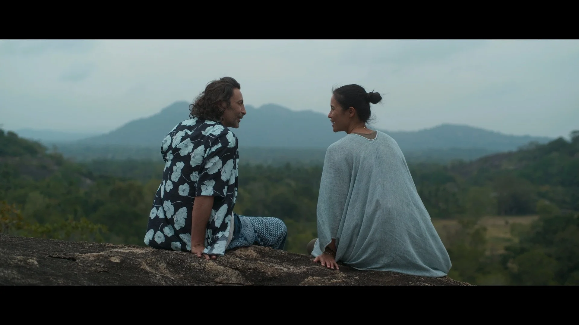 Reset Xuan Lan Filmin Two people sitting on a rock, overlooking a scenic view of mountains and forests, cloudy sky.