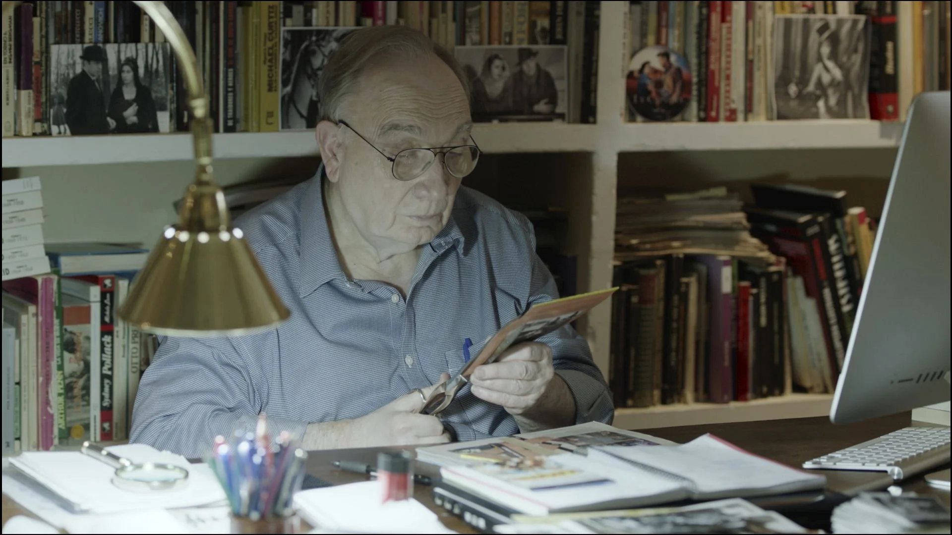 An elderly man with glasses sits at a cluttered desk in a home office, examining a piece of paper. He is surrounded by bookshelves filled with books and photographs. The desk has a computer monitor, keyboard, pens, and various papers. A lamp provides light to the workspace.
