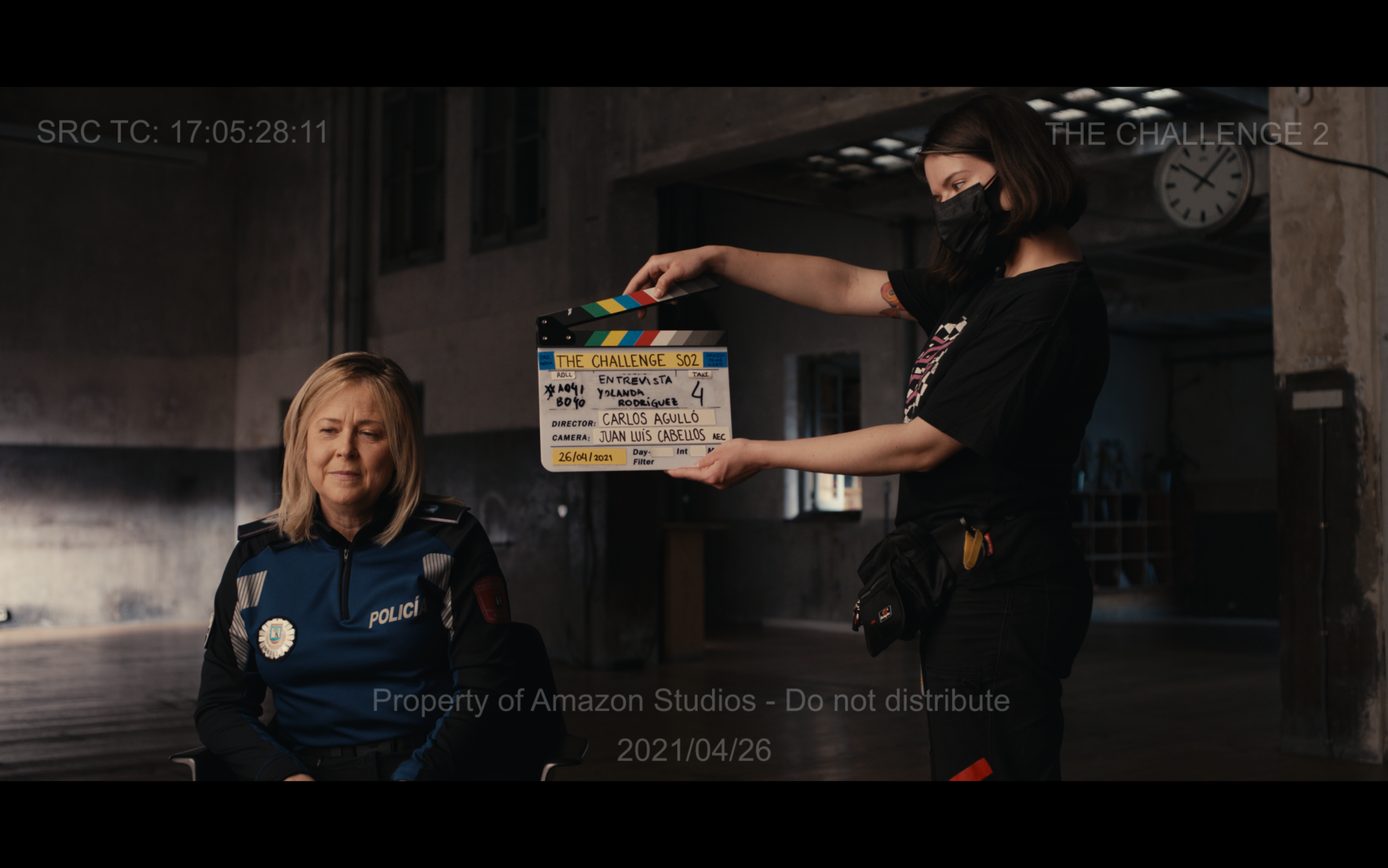 Film scene setup with two individuals, including a woman wearing a "Polići" uniform, and a crew member holding a clapperboard for "The Challenge S02."