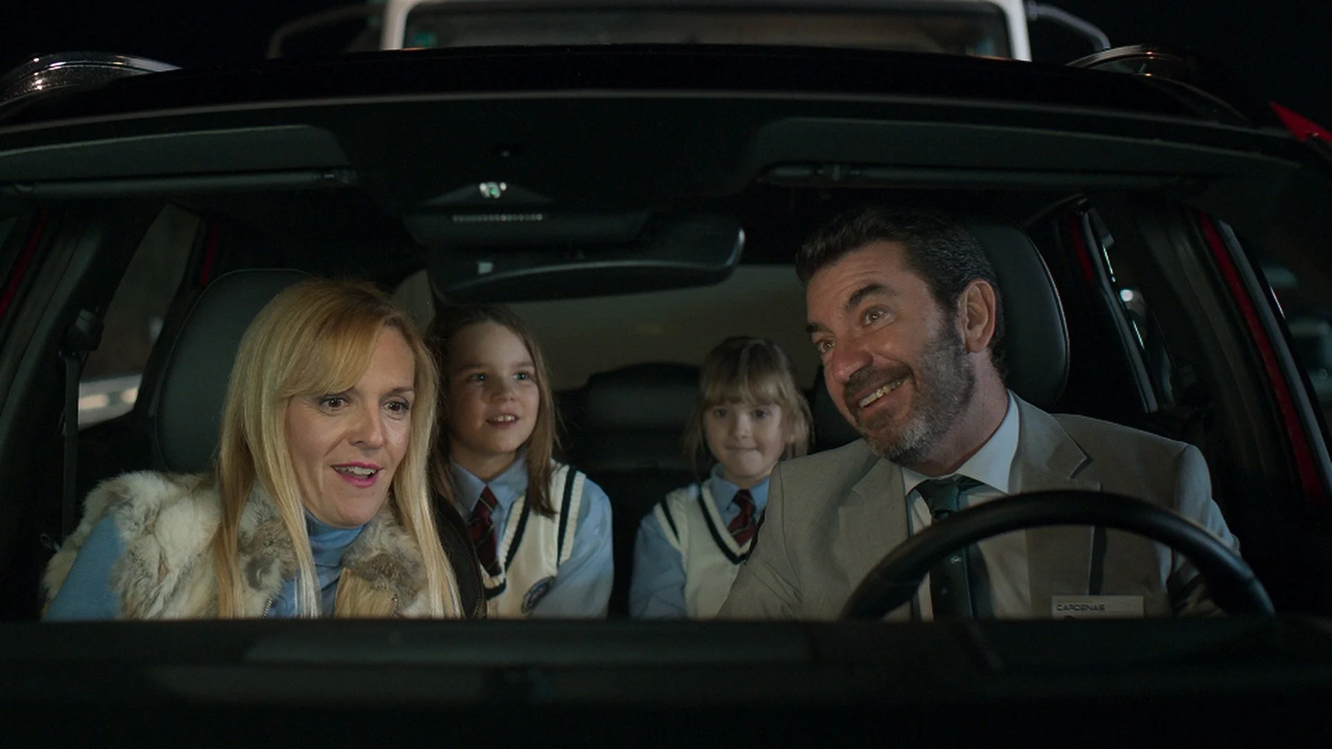 A family sitting in a car, two adults in the front seats and two children in the back wearing school uniforms with ties.
