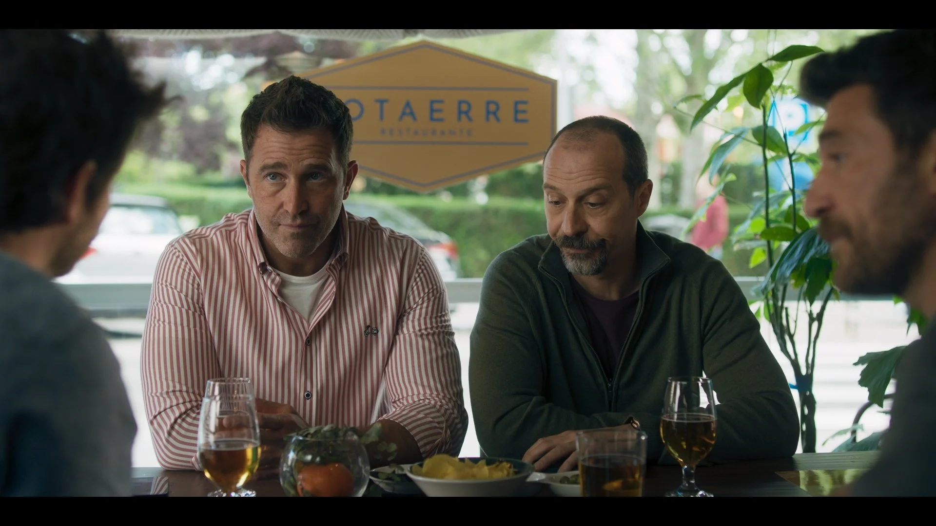 Four men sitting at a restaurant table with drinks, engaged in conversation, with a sign saying 'Restaurante' visible in the background and plants around them.